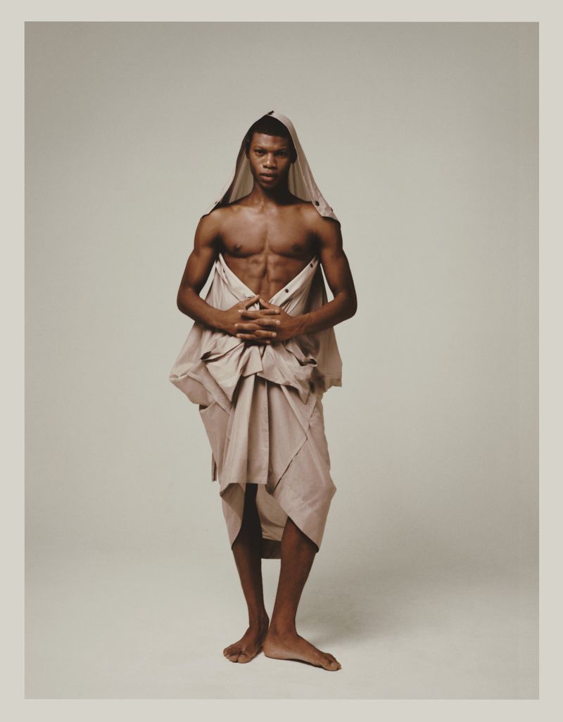 Shirtless Black male model standing barefoot wrapped in draped beige fabric over head and waist against a neutral backdrop.