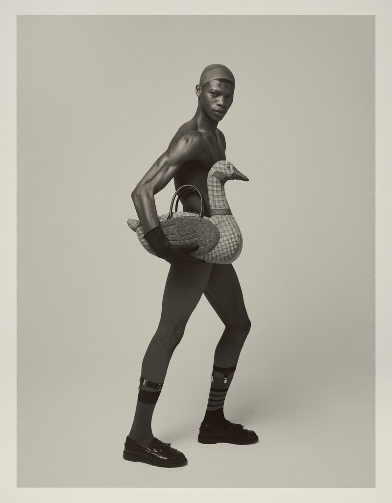 Fashionable, slim, African male model in a cap, socks and loafers, posing while holding a goose-shaped bag against a light-grey background.