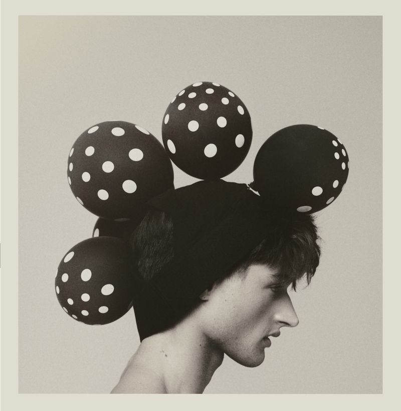 Profile portrait of a young adult male wearing a decorative hat adorned with black polka dot spheres in a studio setting with soft lighting.