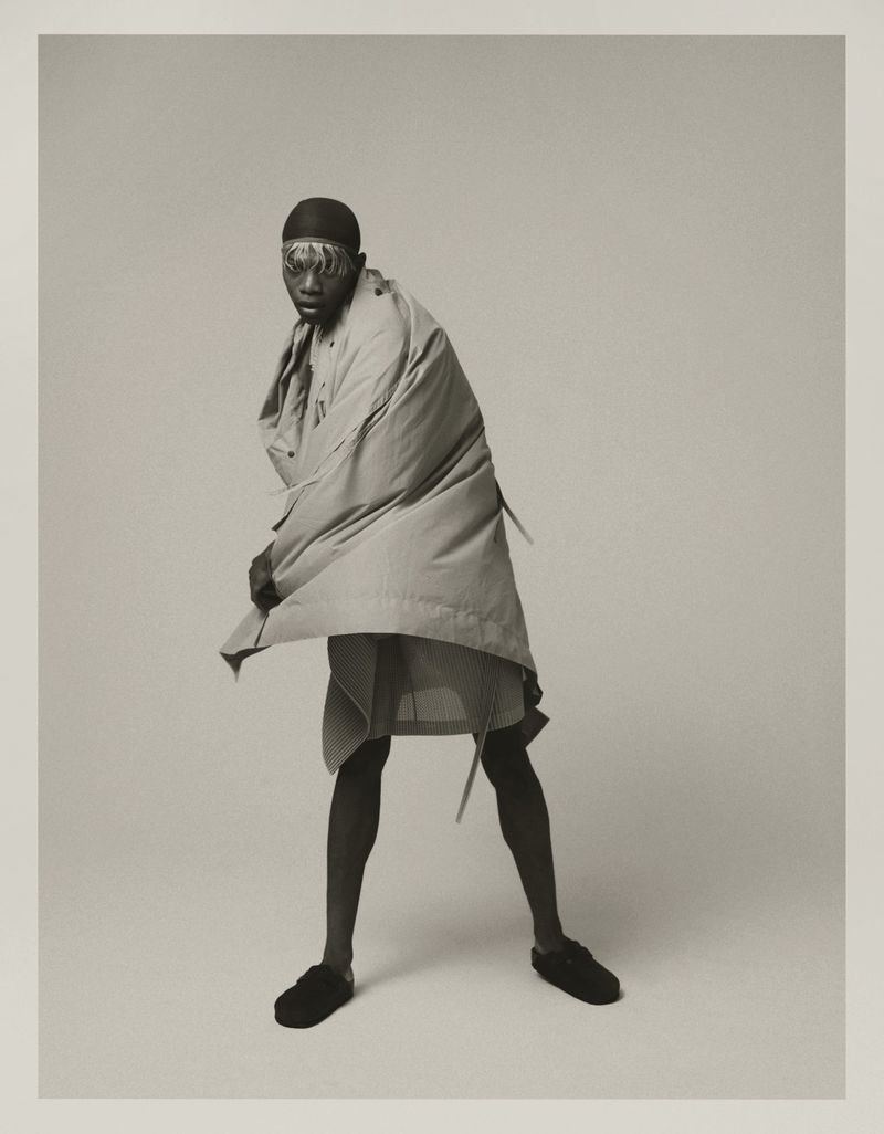 Monochromatic fashion shot of a person posing in an open coat and skirt, with a black cap and slippers, against a light grey background.