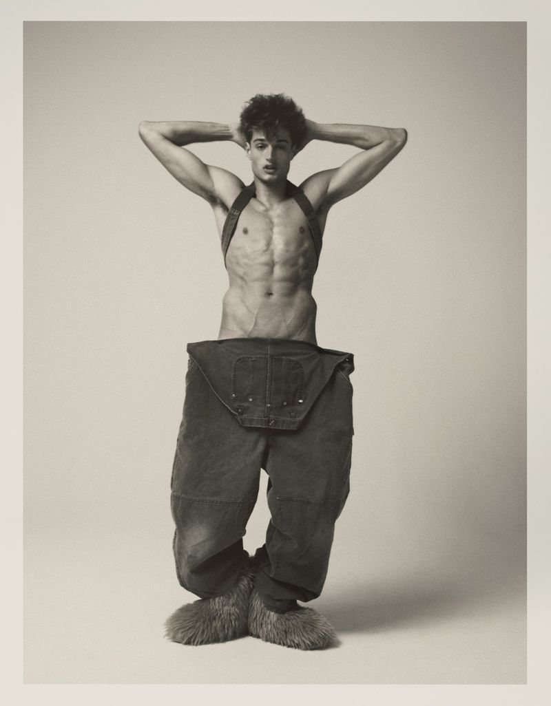Male fashion model poses shirtless in a studio wearing oversized trousers with fur boots, hands behind his head and neutral facial expression.