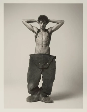 Male fashion model poses shirtless in a studio wearing oversized trousers with fur boots, hands behind his head and neutral facial expression.