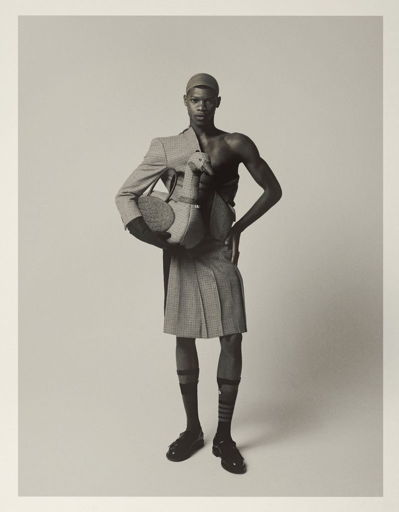 Monochrome studio shot of a Black man wearing a kilt, blazer, and cap holding a stuffed toy goose against a neutral backdrop.