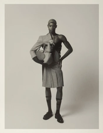 Monochrome studio shot of a Black man wearing a kilt, blazer, and cap holding a stuffed toy goose against a neutral backdrop.