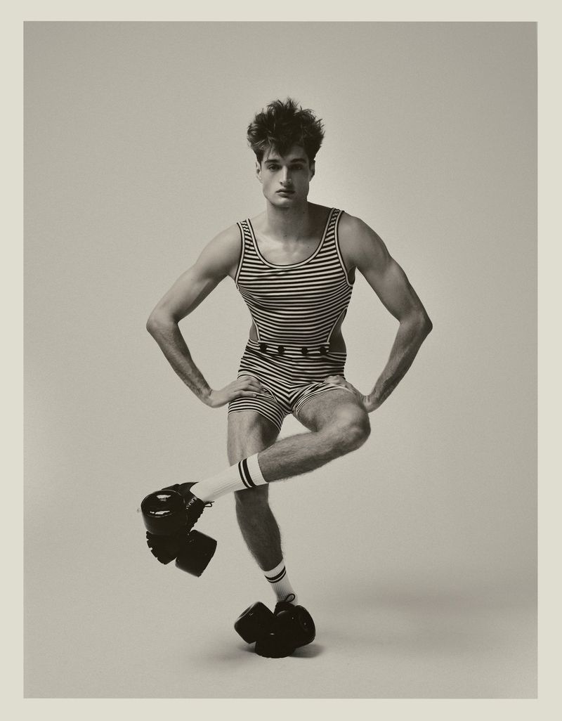 Monochrome studio portrait of a young adult male in vintage swimwear, balancing on one foot with a serious expression.