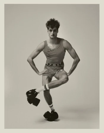Monochrome studio portrait of a young adult male in vintage swimwear, balancing on one foot with a serious expression.