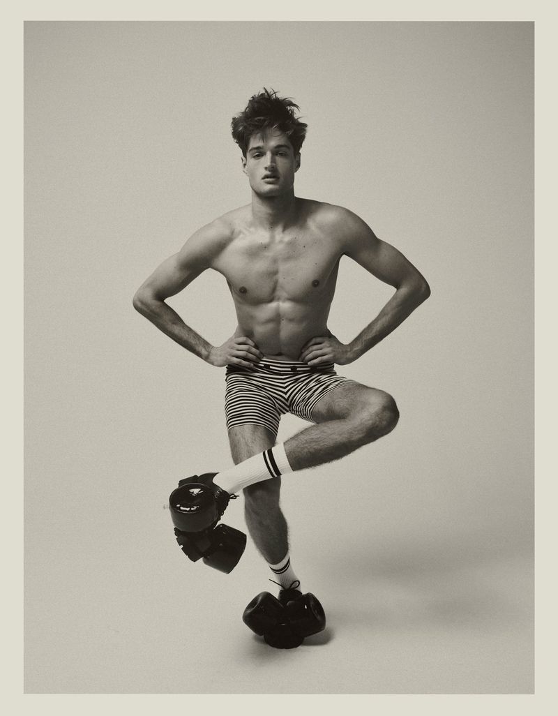 Male fitness model strikes a pose with boxing gloves; indoor setting with neutral background, medium shot, showcasing athletic physique and fashion.