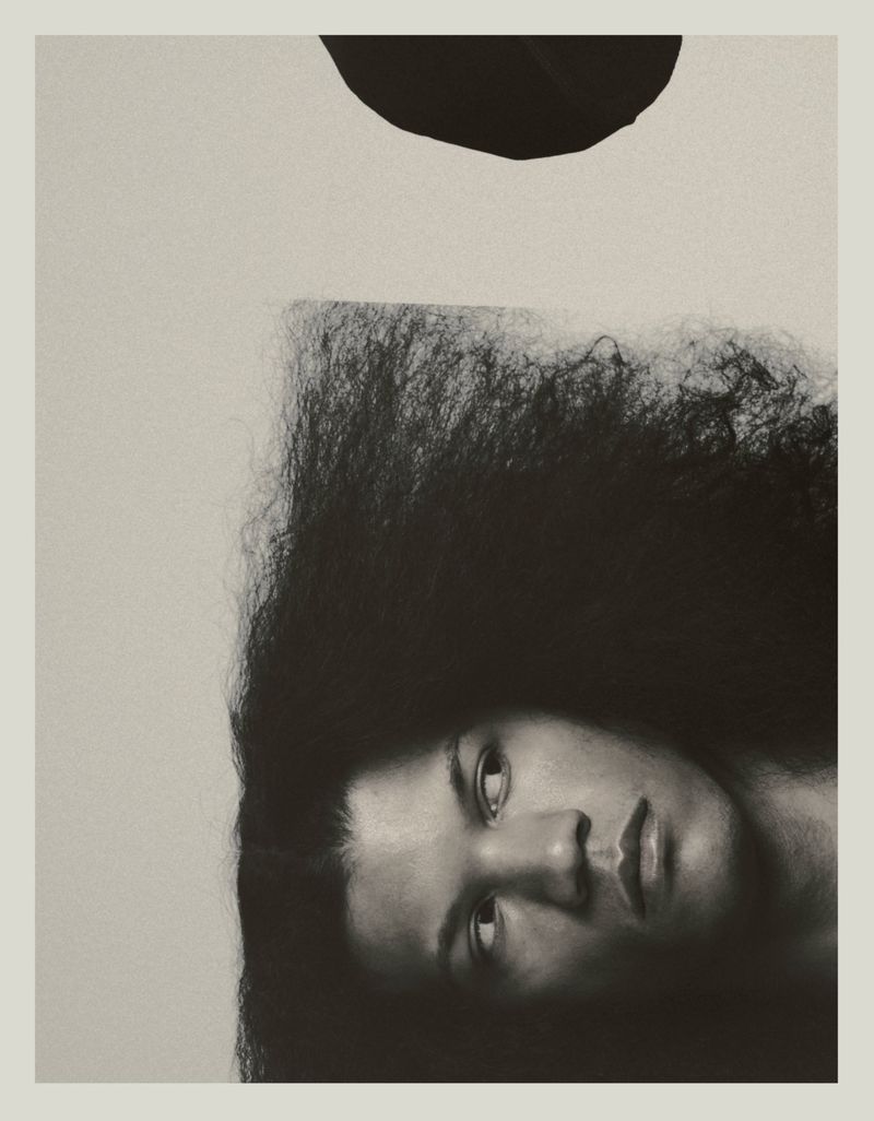 Artistic portrait of a woman with afro hairstyle in black and white with black circle element over her head against a neutral background.