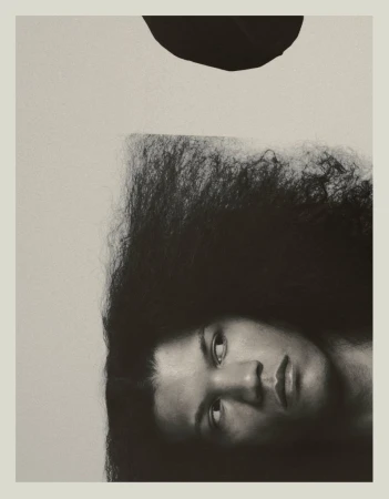 Artistic portrait of a woman with afro hairstyle in black and white with black circle element over her head against a neutral background.