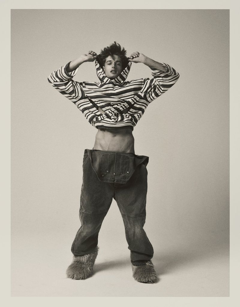 Model Antonio Macek in a striped hoodie and oversized denim overalls with furry boots against a studio backdrop.