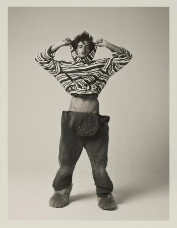 Model Antonio Macek in a striped hoodie and oversized denim overalls with furry boots against a studio backdrop.