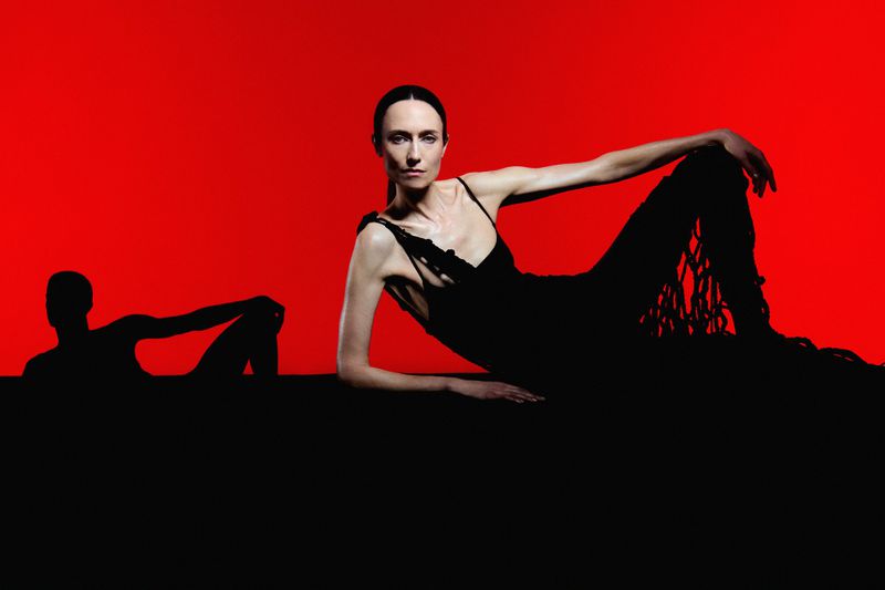 Elegant woman in black gown poses against a vibrant red backdrop, casting a striking silhouette, creating an editorial fashion shot.