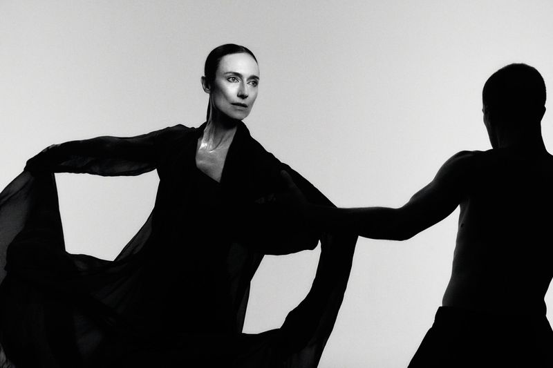 Monochrome photograph of a woman in a dark, flowing garment dancing with a silhouetted man against a light backdrop, showcasing dramatic contrast.