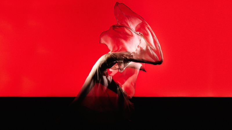 Woman in flowing dress dances in expressive pose against vibrant red background with dark floor in a dramatic performance.