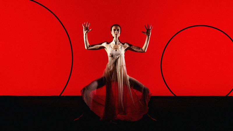 Woman in sheer dress poses in front of a red background with black circle art, showcasing a unique blend of fashion and performance art.