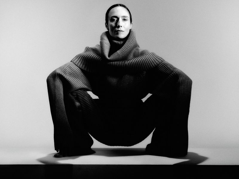 Monochromatic portrait of a woman in a squatting position wearing a gray turtleneck sweater and pants, on a gradient background.