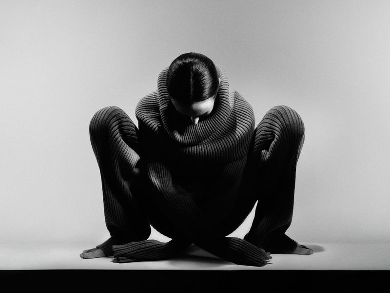 Monochrome shot of an adult woman in a ribbed sweater and pants crouching with crossed legs in a photo studio with a white background.
