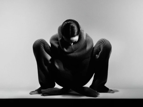 Monochrome shot of an adult woman in a ribbed sweater and pants crouching with crossed legs in a photo studio with a white background.
