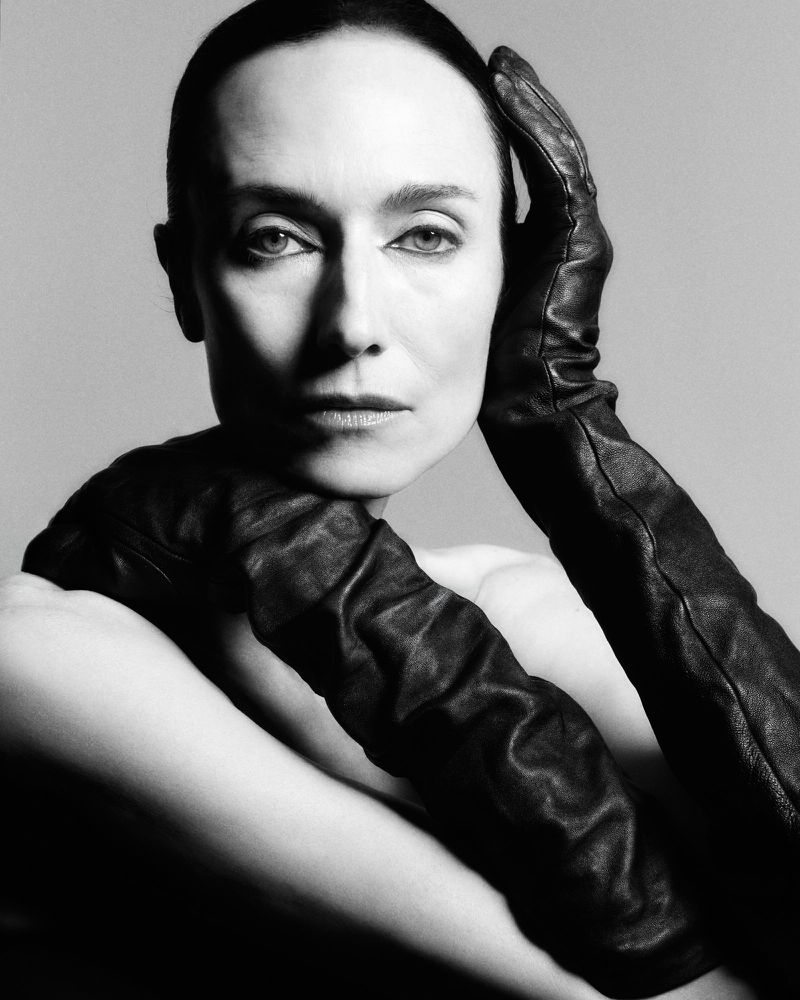 Black and white portrait of a woman with dark hair and eyes, wearing long black leather gloves and posing with her hands near her face.