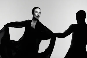 Black and white studio photograph of a woman in flowing dark chiffon fabric extending her arms toward a male dancer silhouette.