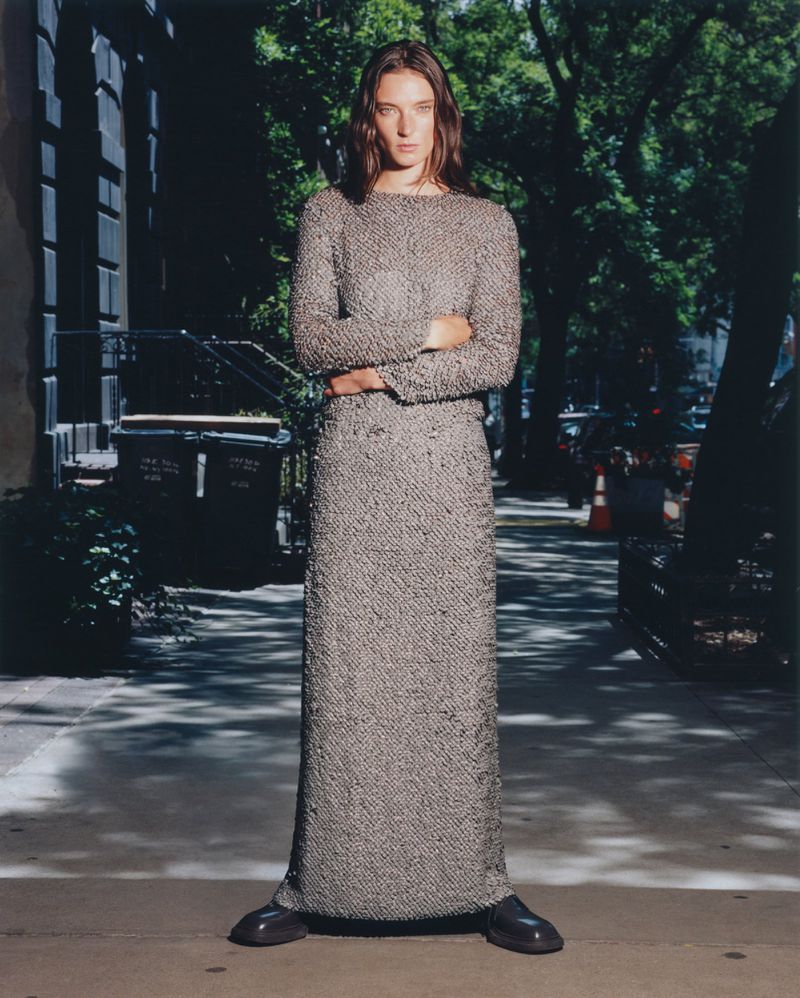Adult woman posing in a long, textured dress with arms crossed in a street setting with trees and townhouses in the background.
