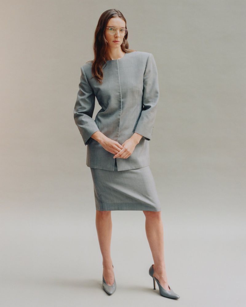 Adult female in a grey skirt suit with matching heels poses against a neutral backdrop in a studio shot. The woman wears glasses.