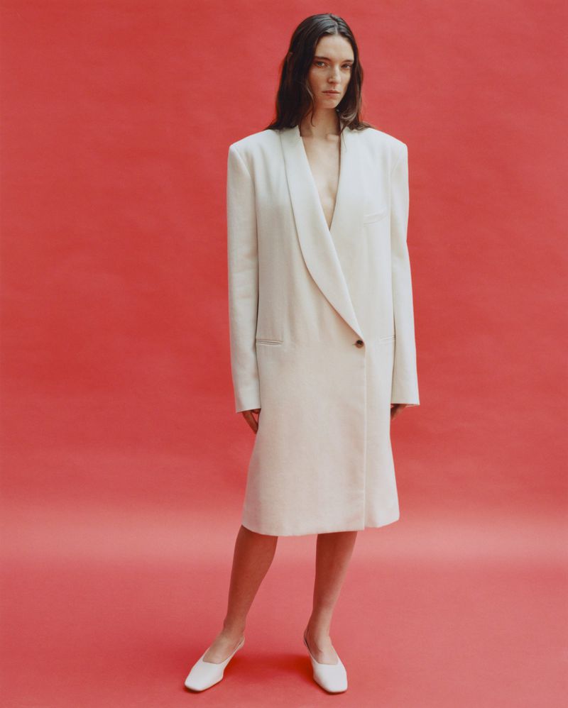 A woman stands in a full shot in a white coat dress with a red studio backdrop, showcasing fashion and simple studio photography.