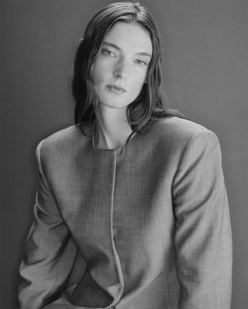 Portrait of an adult female with long brown hair and a neutral expression, wearing a gray blazer against a gray background in black and white.