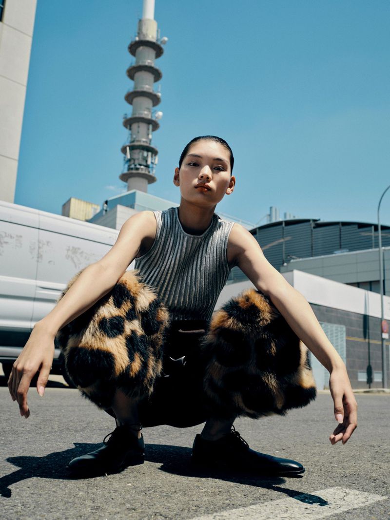 Portrait of an East Asian adult woman crouching on asphalt wearing black shoes and leopard print pants with a gray tank top.