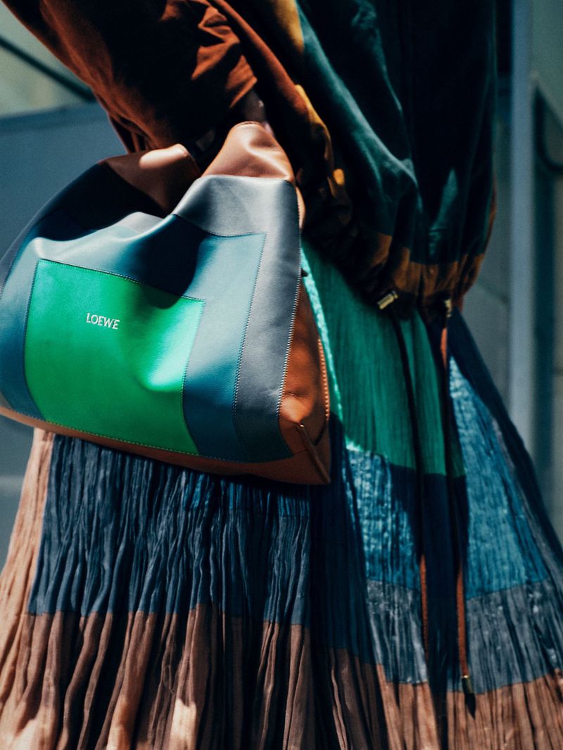 Close-up of a Loewe handbag with green and blue color-blocked panels and a brown leather section, paired with a pleated skirt.