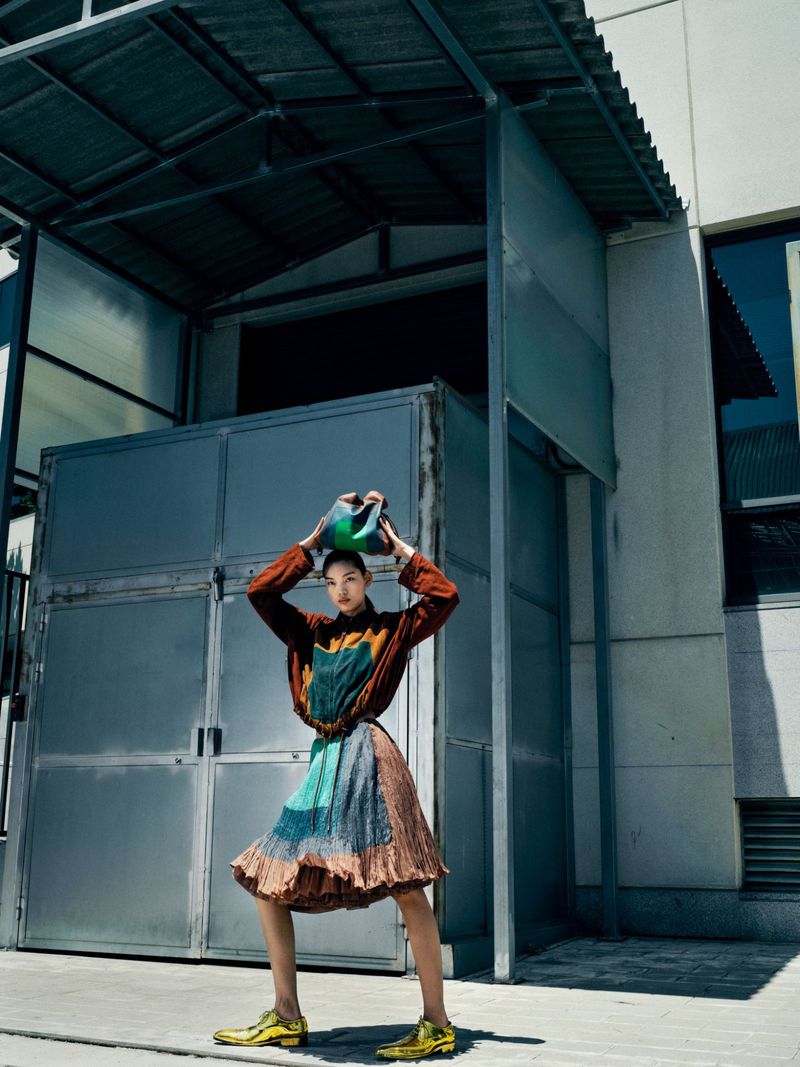 Woman in colorful outfit balances handbag on head in an urban setting with geometric concrete facade on a sunny day.