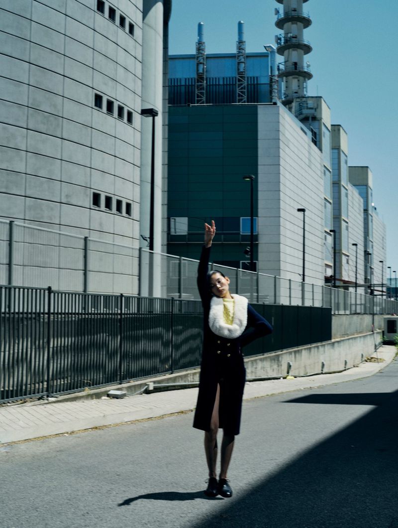 An adult female poses in a coat and skirt on an asphalt road with tall industrial buildings in the background on a bright sunny day.
