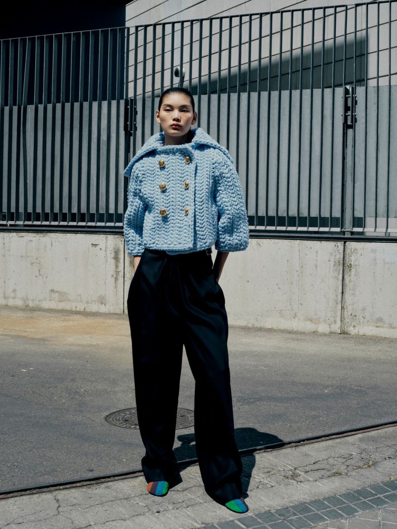Fashionable woman in light blue knitted jacket and dark trousers poses against an urban wall and metal barrier in an outdoor setting.