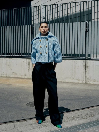 Fashionable woman in light blue knitted jacket and dark trousers poses against an urban wall and metal barrier in an outdoor setting.