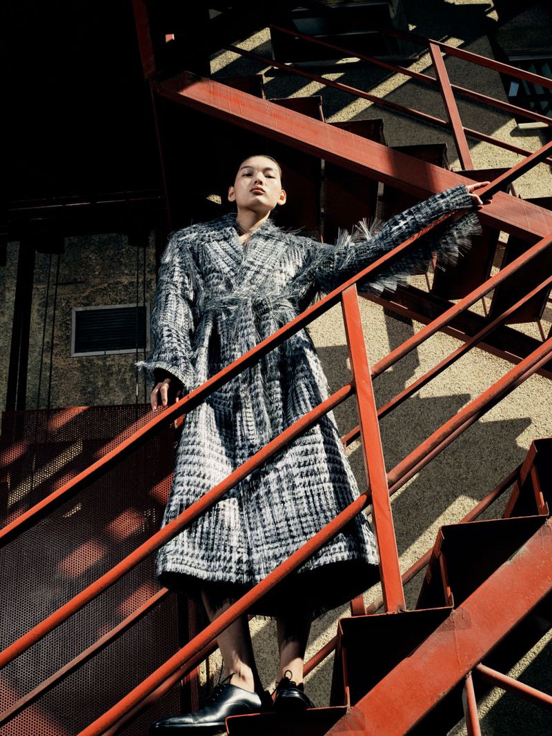 Fashionable adult woman in coat poses on red metal stairway outdoors in daylight, looking up slightly, low angle, monochrome.