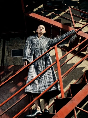Fashionable adult woman in coat poses on red metal stairway outdoors in daylight, looking up slightly, low angle, monochrome.