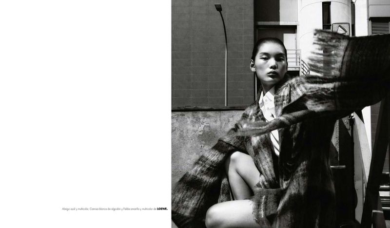 Monochromatic outdoor fashion shot of an Asian adult female model wearing patterned coat and white collared shirt against an urban background.