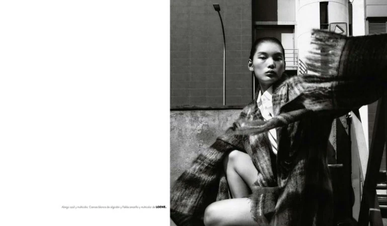 Monochromatic outdoor fashion shot of an Asian adult female model wearing patterned coat and white collared shirt against an urban background.