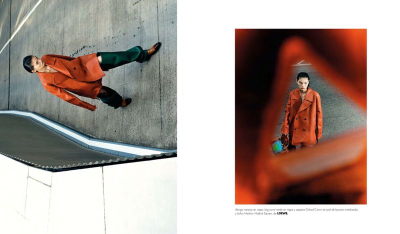 Fashion editorial shot of a model in an orange coat and green pants posing on a concrete structure with bright, contrasting lighting.
