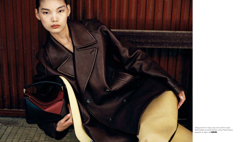 Editorial fashion shot of a woman in a brown leather coat holding a color-blocked handbag against a corrugated background.