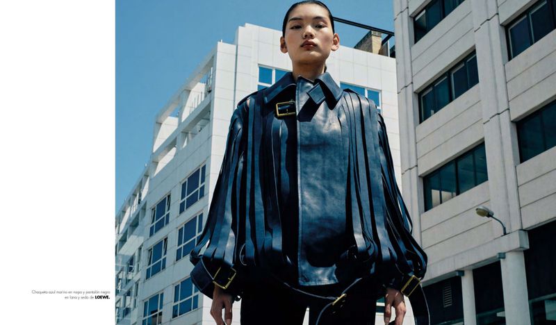 Fashion editorial shot of an Asian model wearing a navy blue leather jacket with fringe and black pants against a neutral urban background.