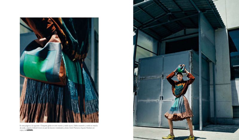 Fashion editorial shot of a model holding a green Loewe handbag above her head, wearing a brown patterned jacket and pleated skirt.