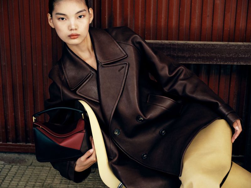 A stylish Asian woman in a brown leather coat poses with a color-blocked handbag against a rusty red metal wall, fashion.