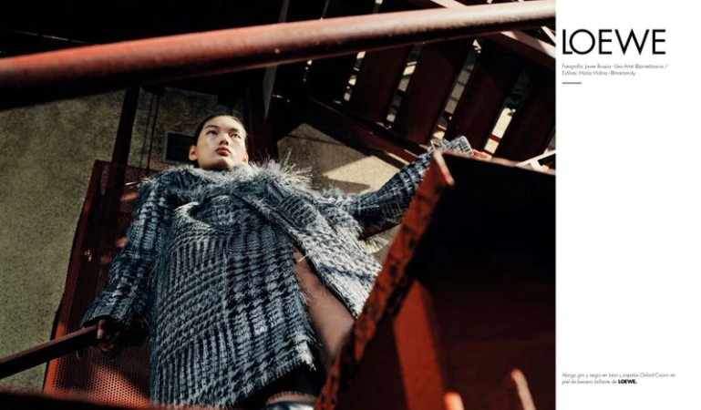 Fashion shot of an Asian female model in a tweed coat with feathered trim posing on metal stairs in an urban setting with overhead lighting.