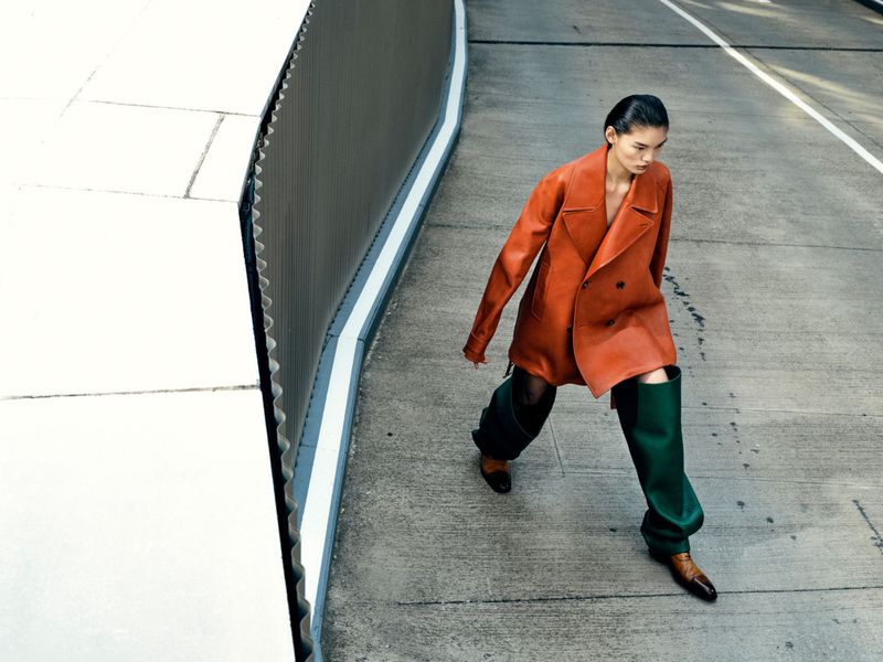 Fashion shot featuring an adult woman in an orange coat and green thigh-high boots walking on a sidewalk with a modern building in the background.