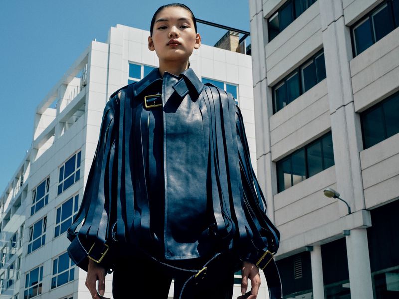 Fashion shot of a stylish adult woman with dark hair wearing a blue fringed leather jacket, photographed outdoors with white buildings.