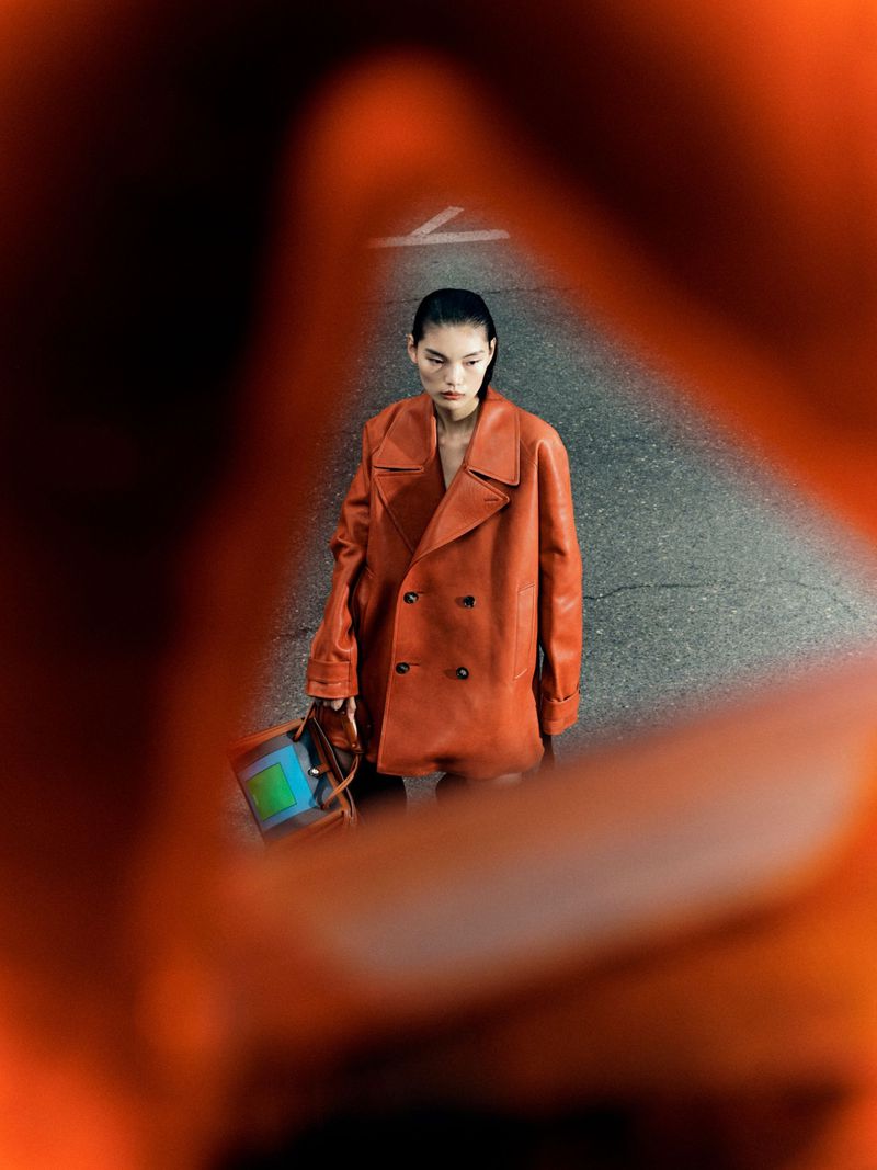 Woman in a double-breasted orange coat stands on pavement, holding a color-block purse. Overhead view through an orange, blurred foreground.