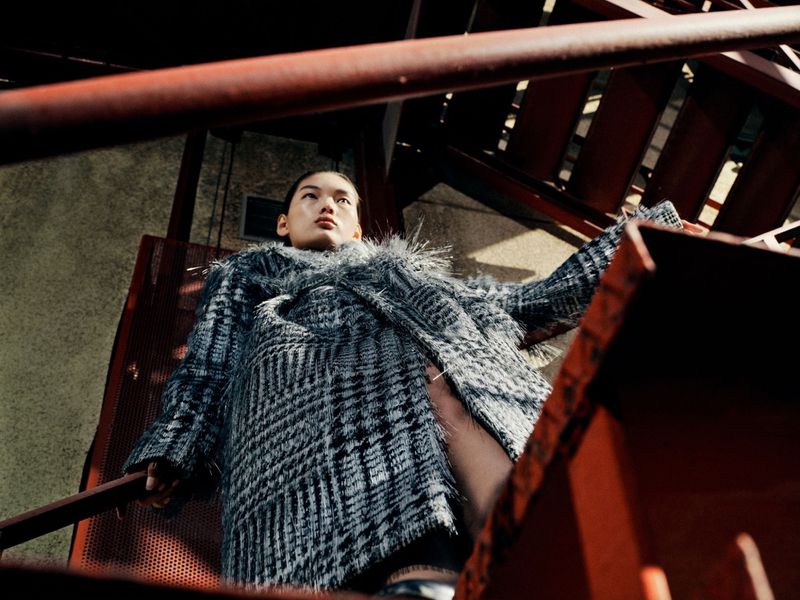 Fashionable young Asian woman in stylish patterned overcoat poses confidently on metal stairs in industrial location with an upward angle.