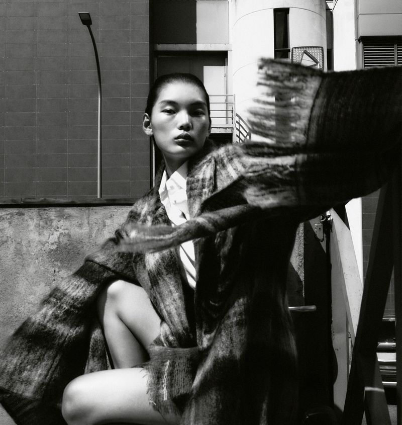 Monochrome portrait of Asian female model in checked coat, seated outdoors against industrial background, evoking high fashion.