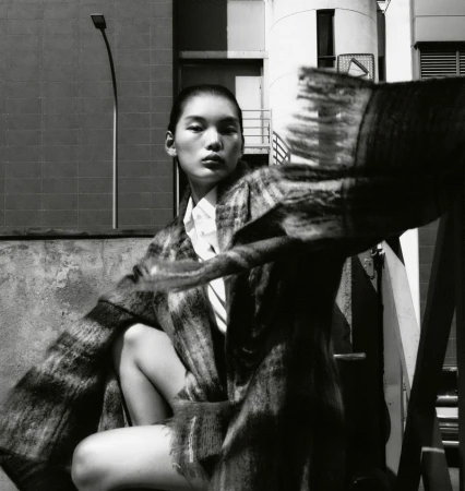 Monochrome portrait of Asian female model in checked coat, seated outdoors against industrial background, evoking high fashion.